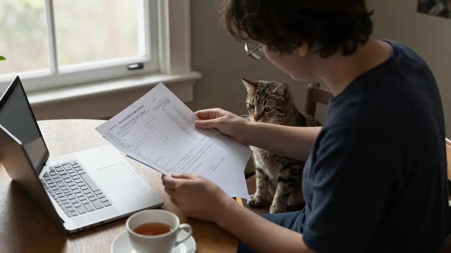 A pet owner at a dining table reading a printed itemised invoice beside a laptop and a cup of tea