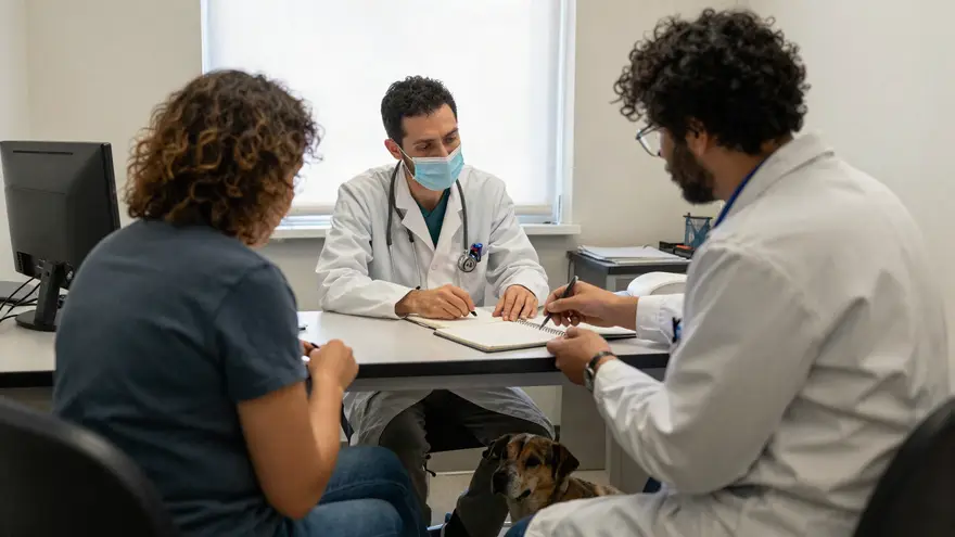 An owner talking with a vet across a consult desk with an open notebook and an old dog resting at their feet